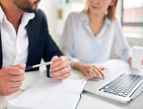 Hands of businessman with pen over open notebook during interaction with colleague